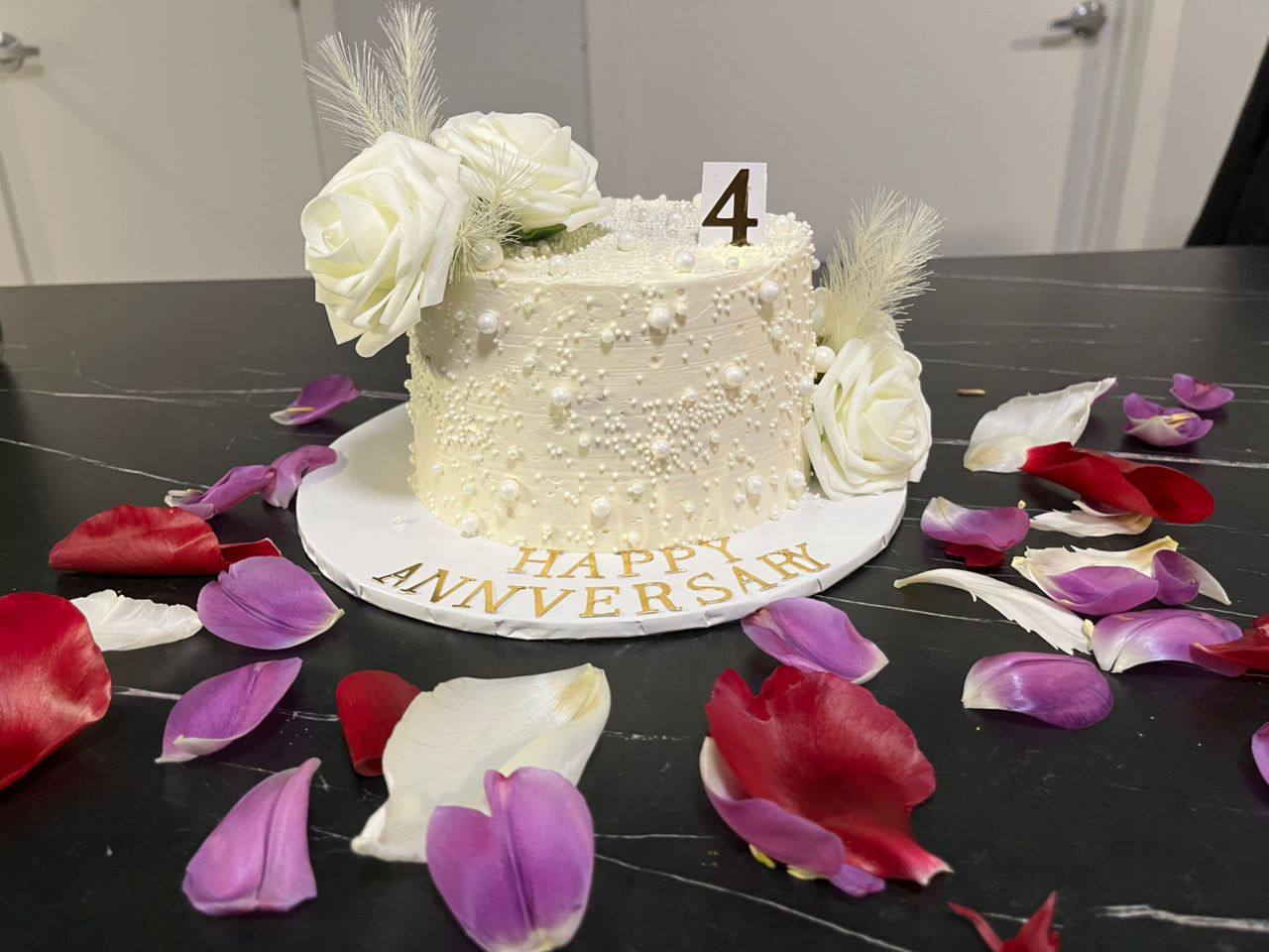 Ivory pearl-beaded anniversary cake with white roses and feathers, a gold 4 topper, 'Happy Anniversary', styled with rose petals