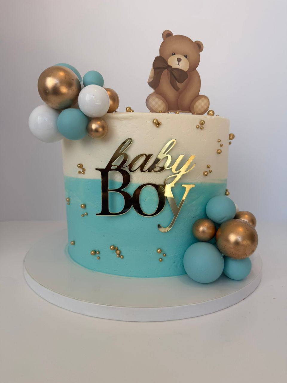 Half white, half teal baby boy cake with gold balloon cluster and teddy bear topper