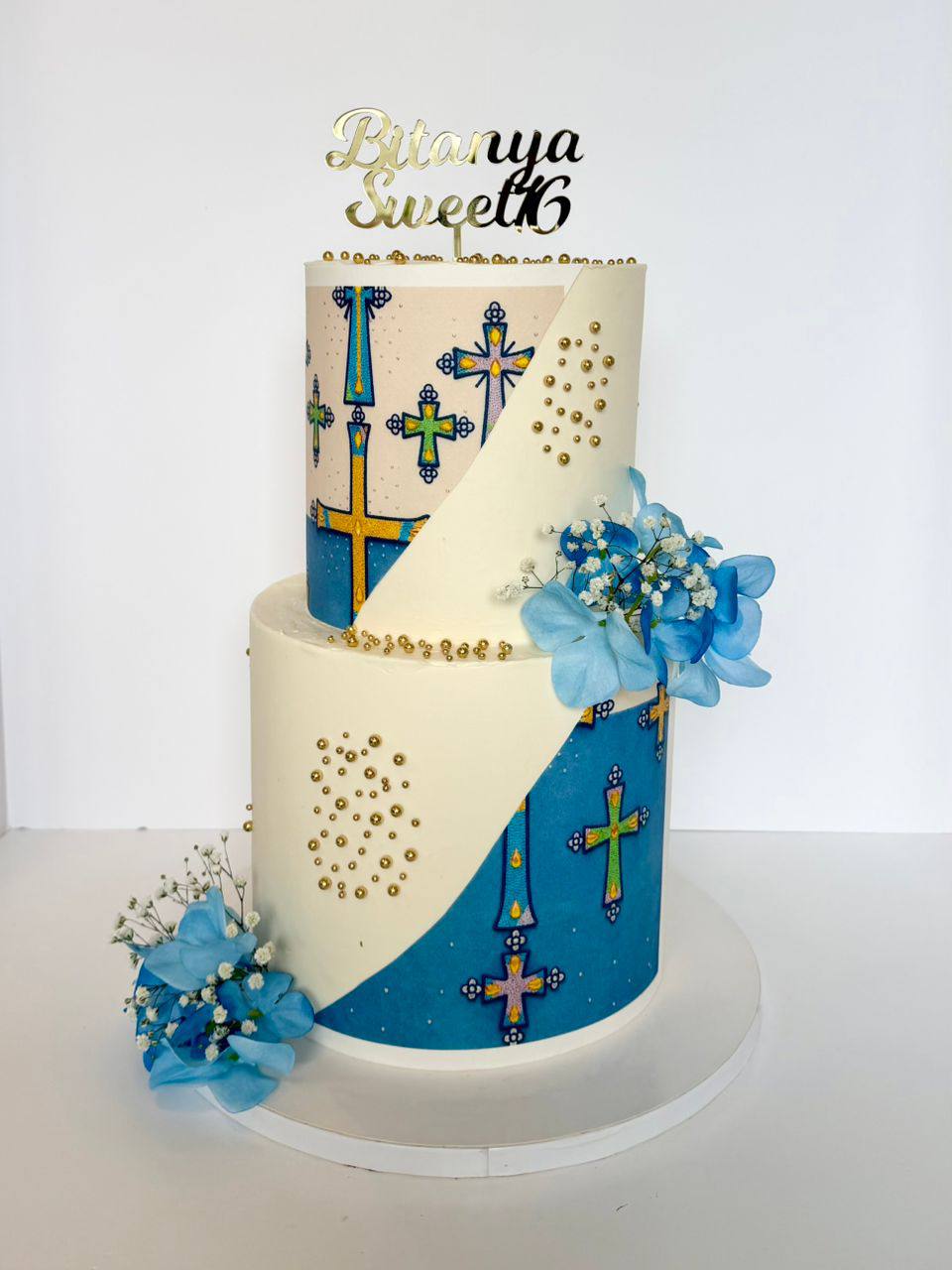 Two-tier white cake with blue stained-glass cross panels, gold pearl clusters, and blue florals, 'Bitanya Sweet 16' topper
