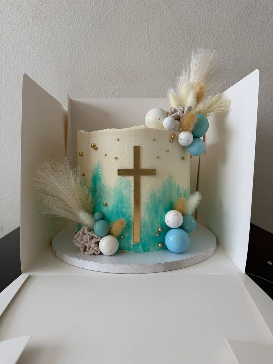 Ivory and teal watercolor christening cake with a gold cross, blue and white spheres, gold pearls, and dried pampas grass