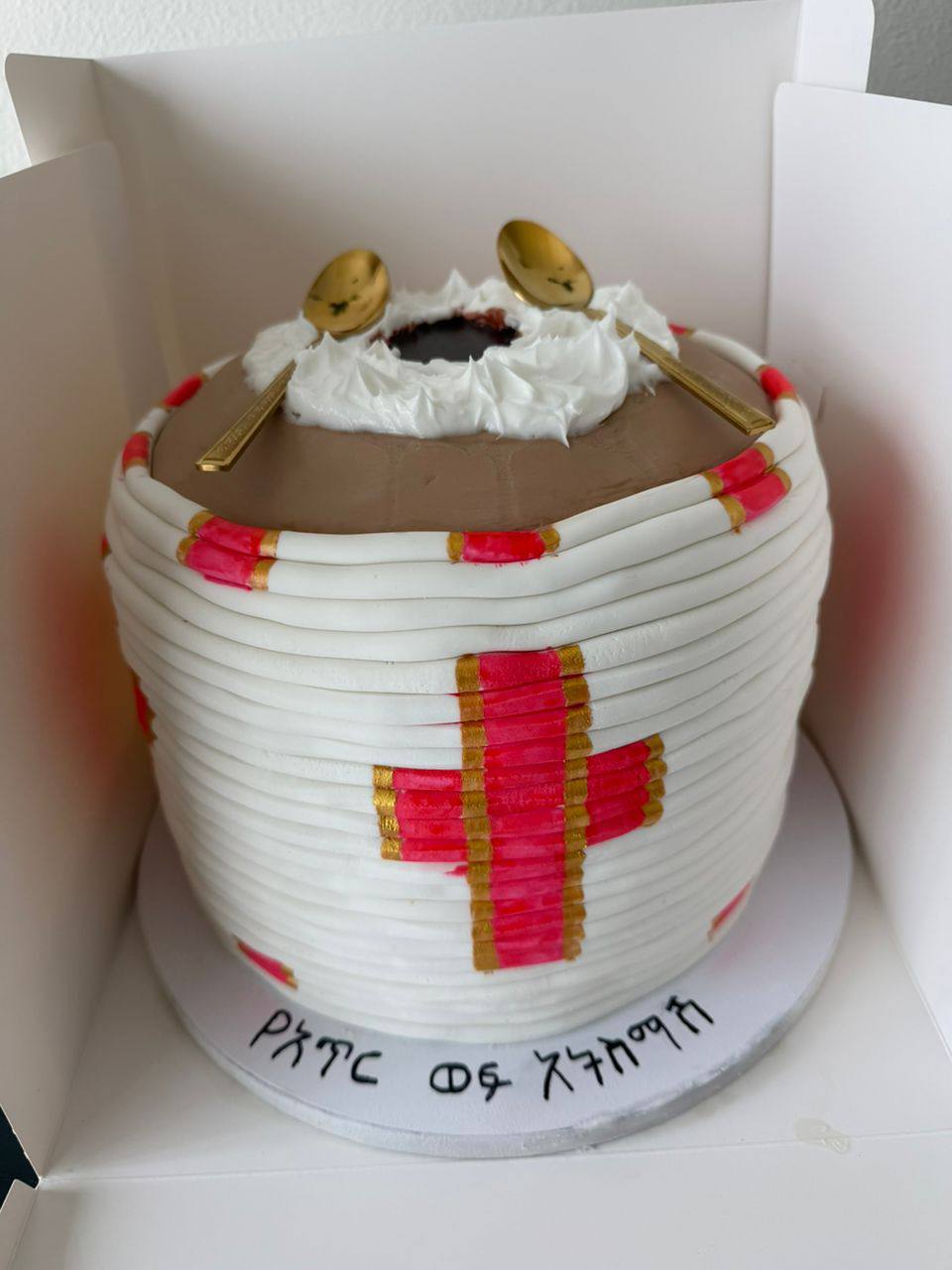 Woven fondant coffee-ceremony cake with a red and gold cross, whipped coffee top, and gold spoon toppers, Amharic lettering