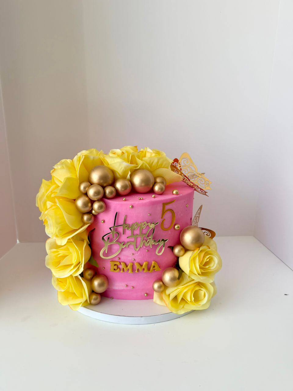 Pink cake framed with yellow roses, gold spheres, and a gold butterfly, 'Happy Birthday Emma' with a gold 5