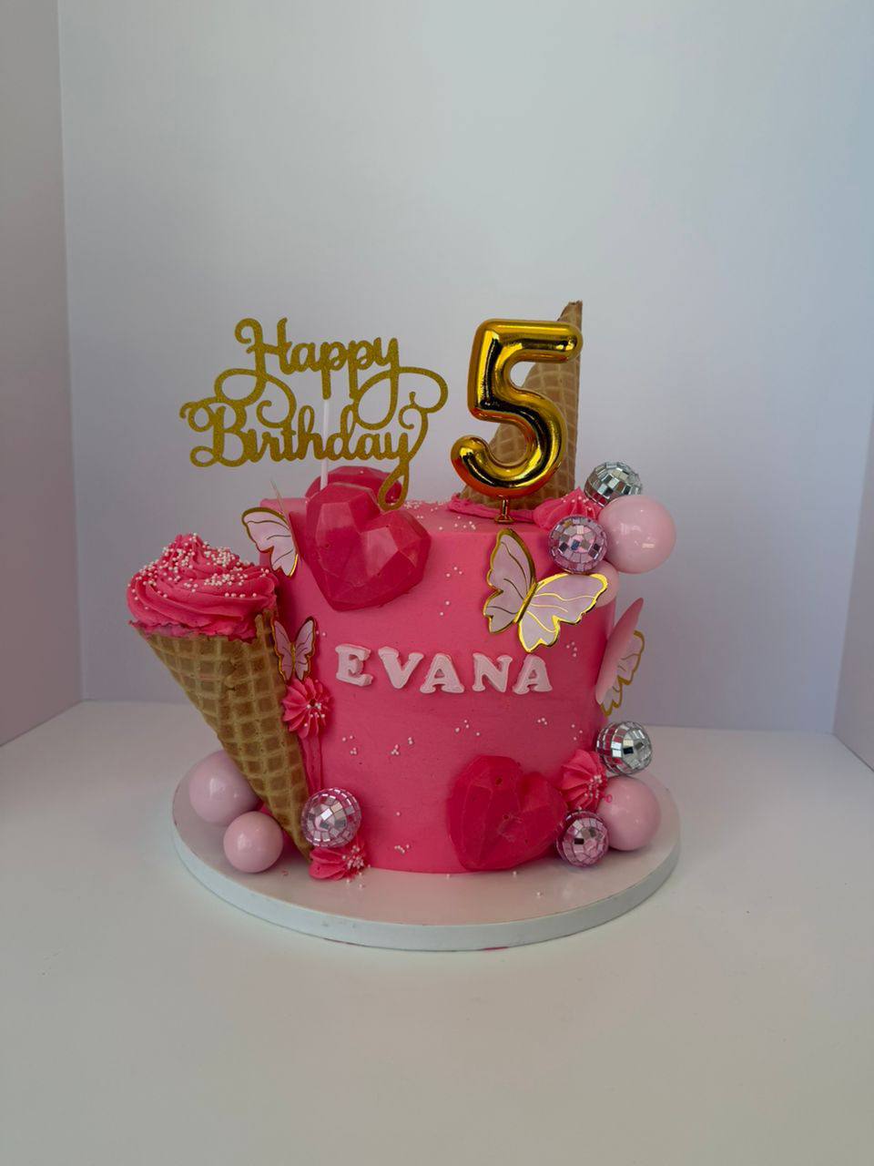 Hot pink cake with an ice cream cone, gold butterflies, disco balls, and a gold '5' topper, name Evana
