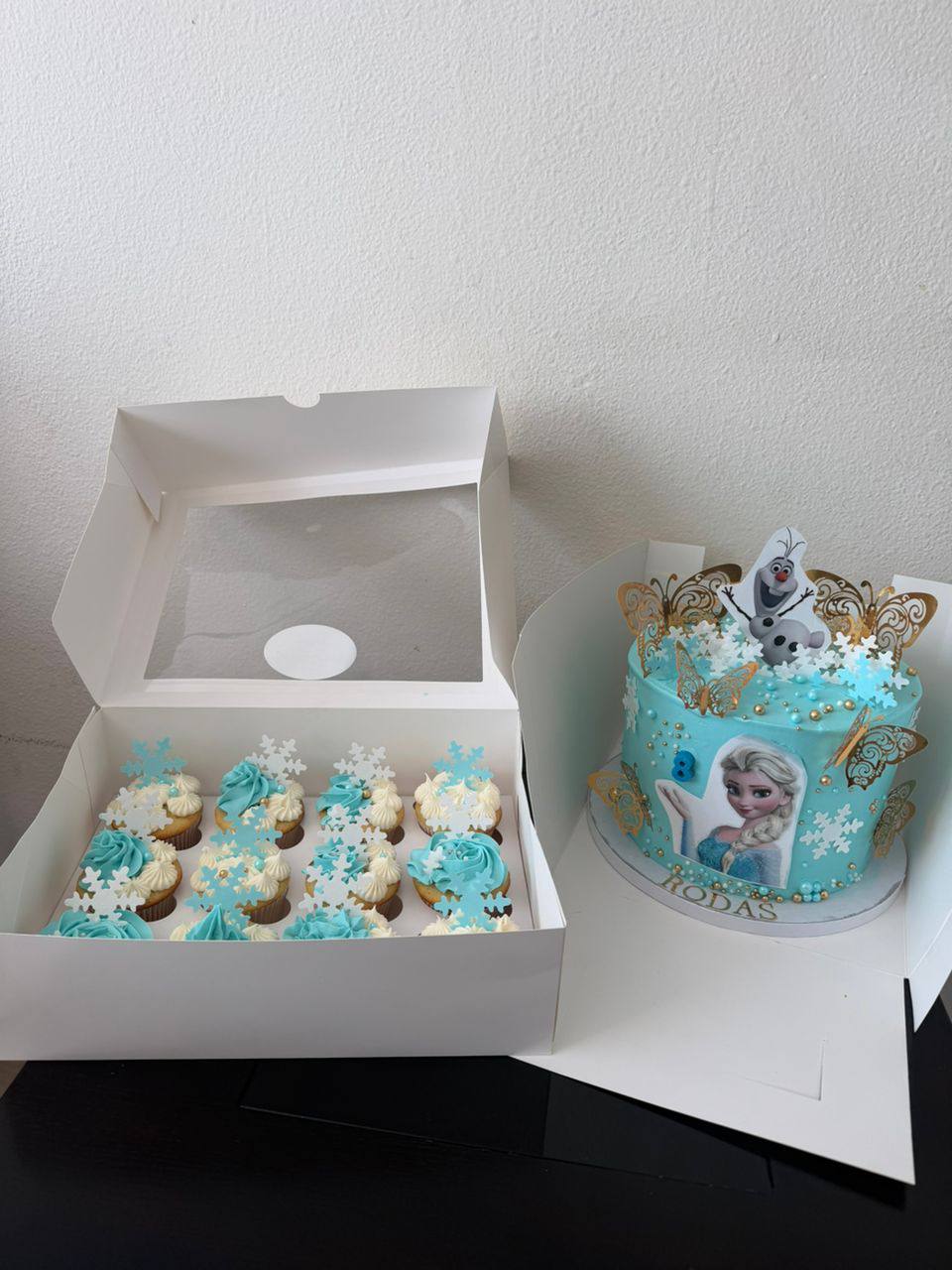 Boxed set of teal and white Frozen-themed cupcakes with snowflake toppers beside a matching cake