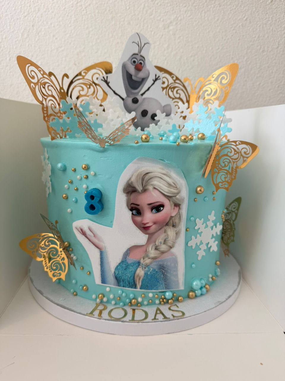 Icy blue Frozen-themed cake with printed Elsa image, fondant Olaf, and gold butterflies, name Rodas