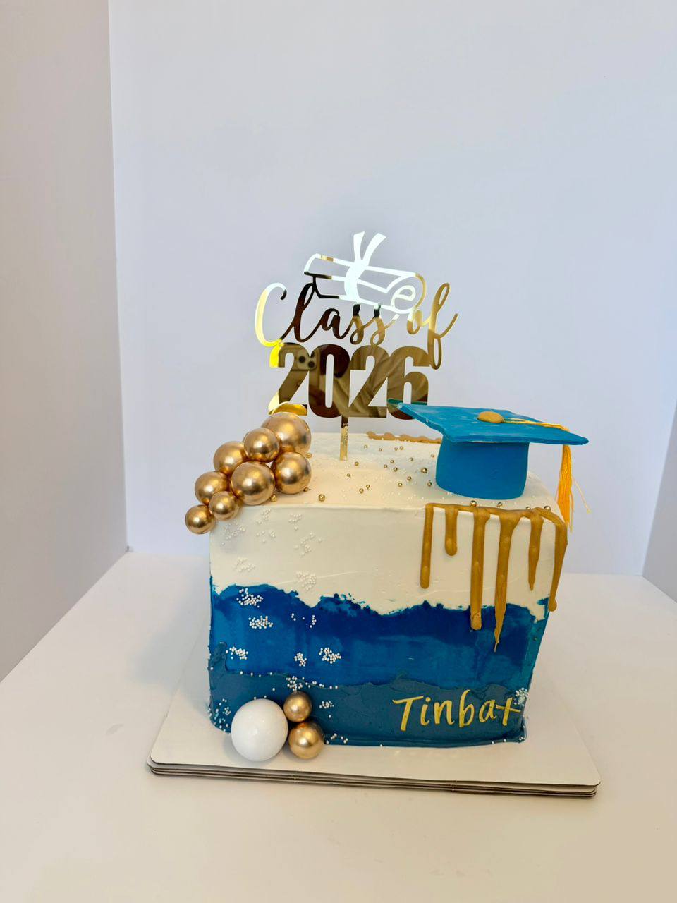 Blue and white watercolor graduation cake with a fondant cap, gold drip, gold spheres, and 'Class of 2026 Tinbat' topper