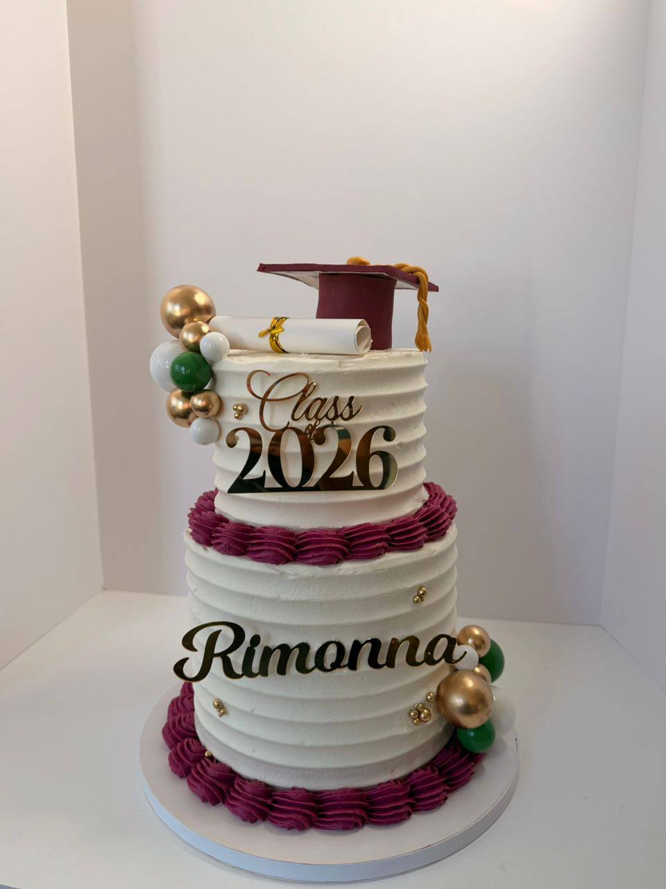 Two-tier white and maroon graduation cake with a fondant cap, diploma, gold and green spheres, 'Class of 2026 Rimonna'