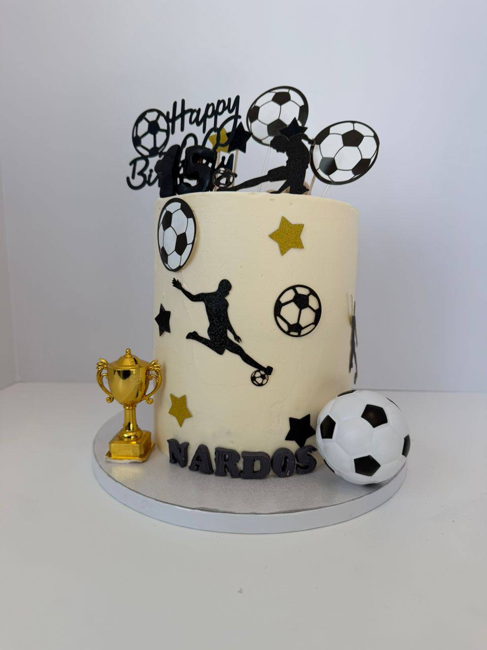 Tall ivory soccer cake with player silhouettes, a gold trophy, fondant soccer ball, and gold stars, name Nardos