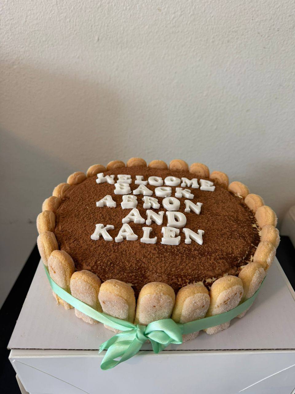 Classic tiramisu cake dusted with cocoa, ringed with ladyfingers and a mint ribbon, 'Welcome Back Abron and Kalen' lettering