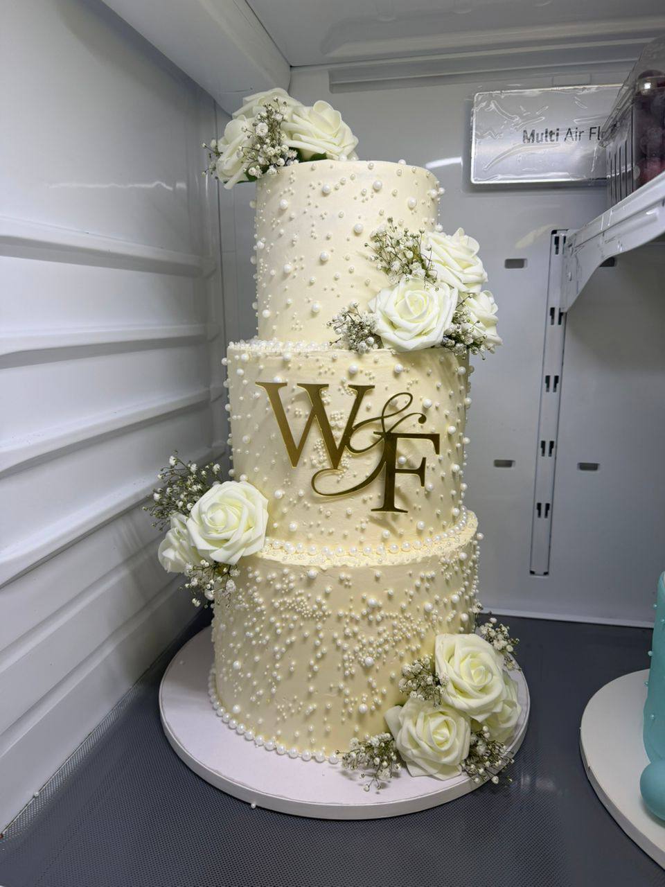 Three-tier ivory wedding cake with pearl beading, white roses, and gold W&F monogram